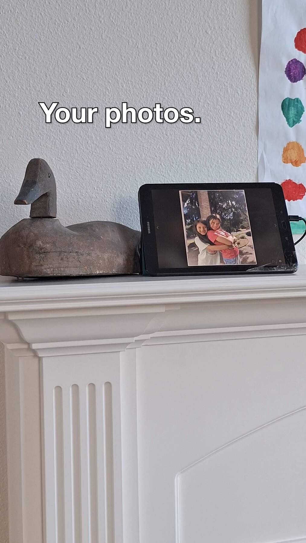 A tablet on a shelf displaying a photo of two daughters hugging.